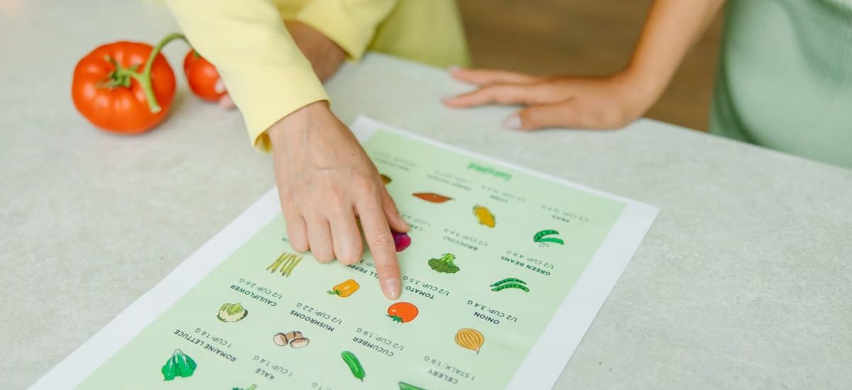Close-up of hands pointing to a vegetable nutrition chart with fresh tomatoes on the table.
