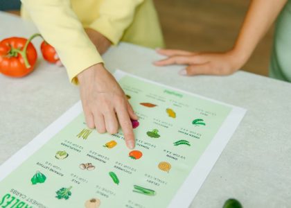 Close-up of hands pointing to a vegetable nutrition chart with fresh tomatoes on the table.